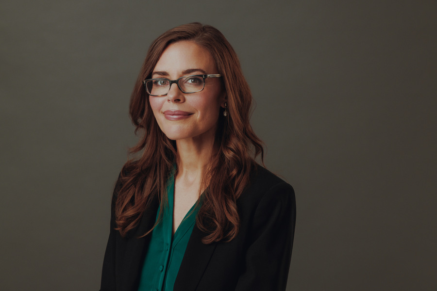 Woman with brown hair and glasses in a green button-down and dark blazer, body at an angle, looking into the camera and smiling.
