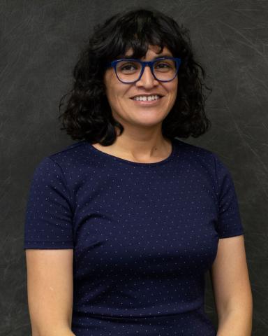 A woman with glasses and shoulder-length dark hair facing the camera and smiling against a heather grey background.