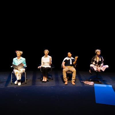 Several actors in chairs, facing the audience, in various poses.