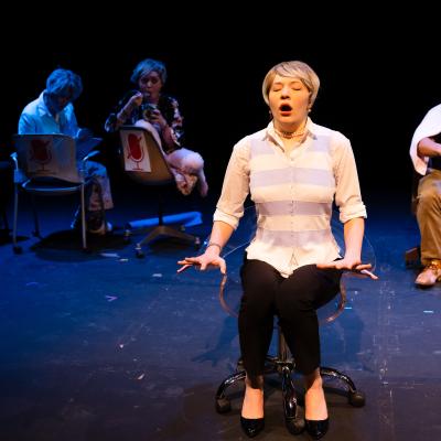 An actress in the foreground, shocked expression, other actors in chairs behind.