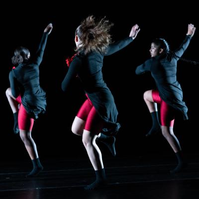 Three dancers with dark hair, wearing dark tops and red shorts, in unified motion