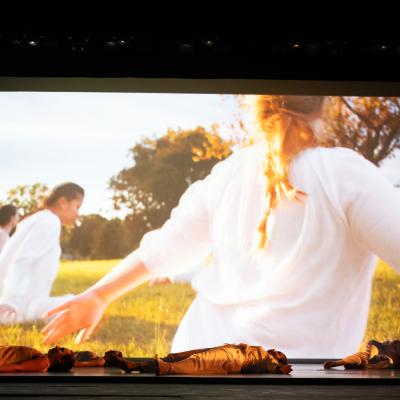 dancers lying on stage, film on screen behind of dancers outdoors, seated