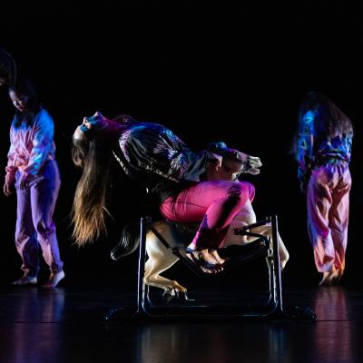 five dancers in colorful outfits, four in the background in various poses, one on a rocking horse, in motion