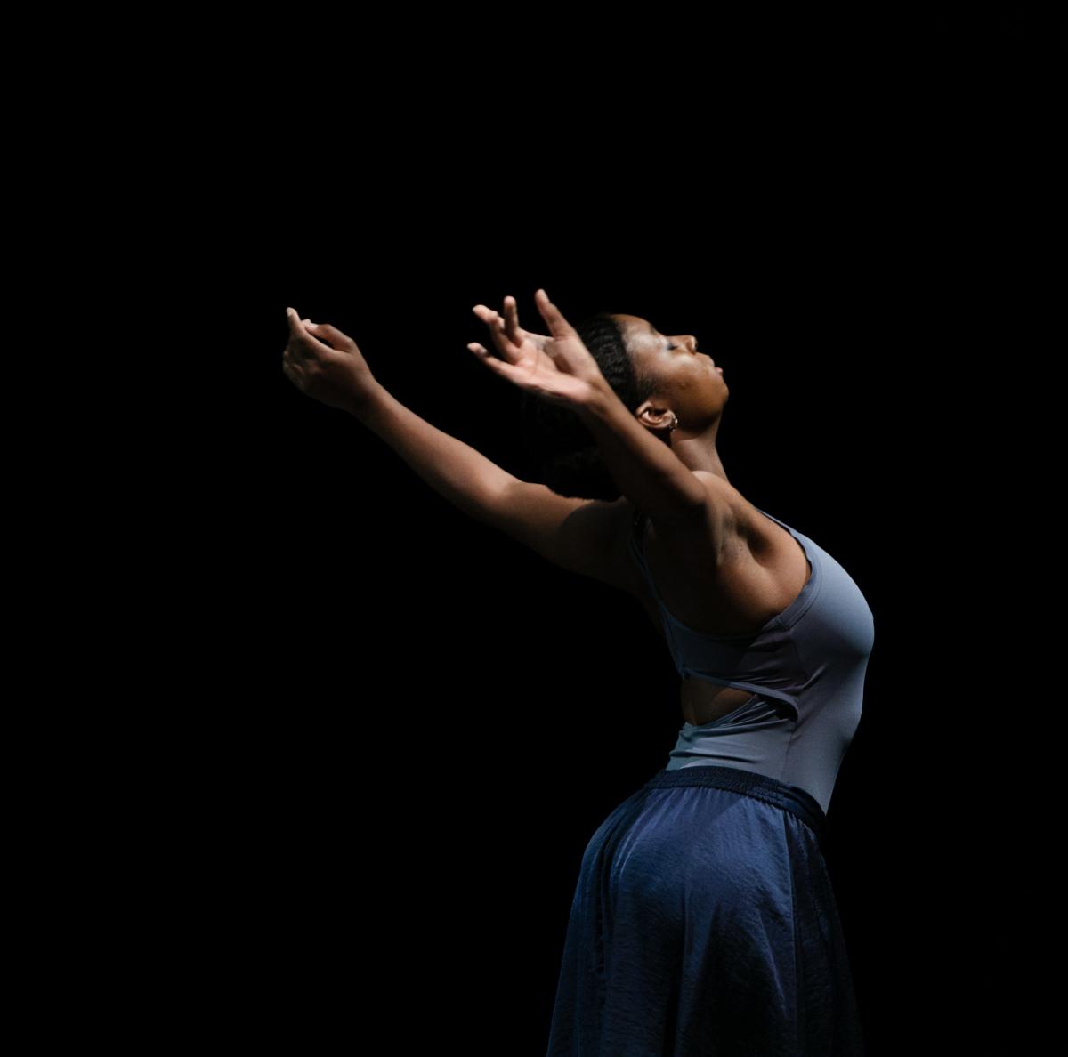 Dancer in blue, in motion at right side of image, back arched, arms outstretched up and behind,
