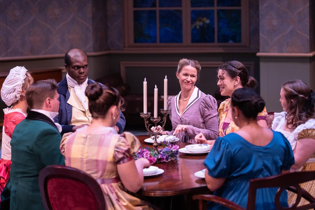 Various actors, in period dress, around a large dining table.