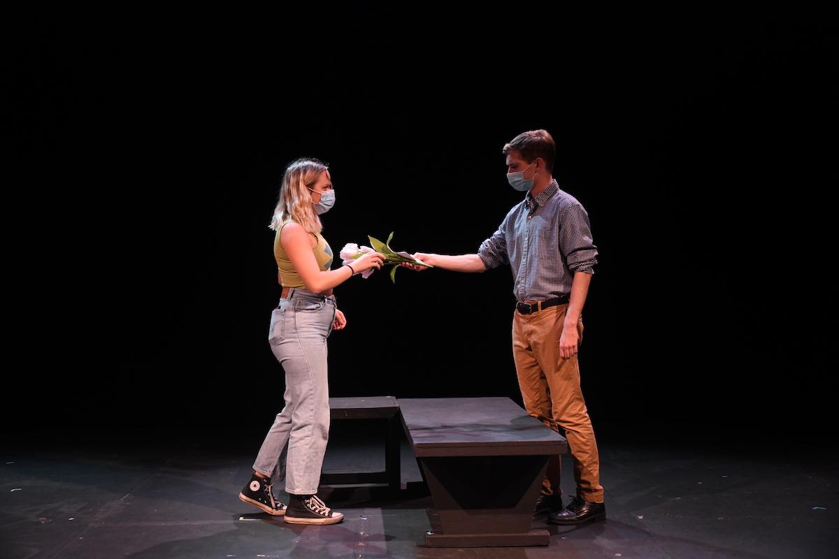 Two actors facing each other with a low table or bench between them, one handing flowers to the other.