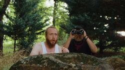 Two figures in a forest, crouching behind a large log, one with binoculars.