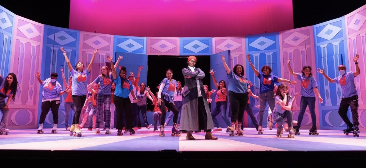 End of musical number pose, young woman dressed as a man in trench coat and hat, with a large chorus in various poses behind, pink and blue set as background.