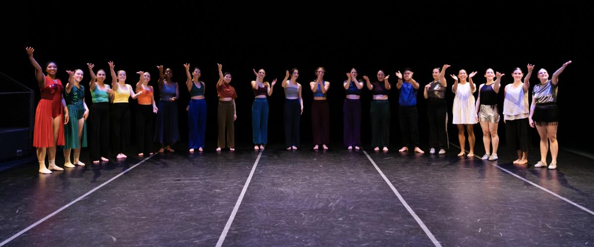 19 dancers with costumes of various colors spanning the rainbow, in a semi-circle for curtain call, dark background