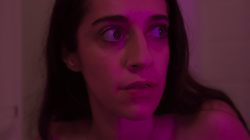 Face of a young woman with dark hair and a startled or anxious expression, reddish lighting