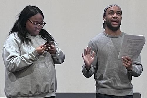 Two figures onstage, one speaking with arms partially raised, the other looking on.