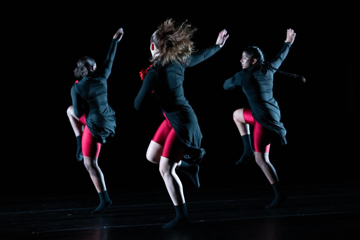 Three female dancers in motion, wearing black tops and red shorts.