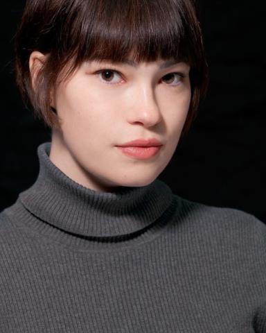 Woman with dark hair and bangs, grey turtleneck, facing the camera at an angle, black background