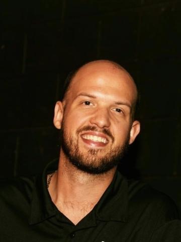 Man facing the camera and smiling, dark facial hair, dark background
